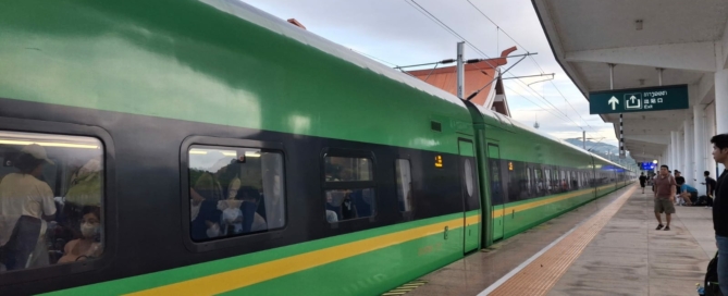 Green and yellow Laos-China Railway trainset at the platform in Luang Prabang station, passengers on board and exit sign in Lao script