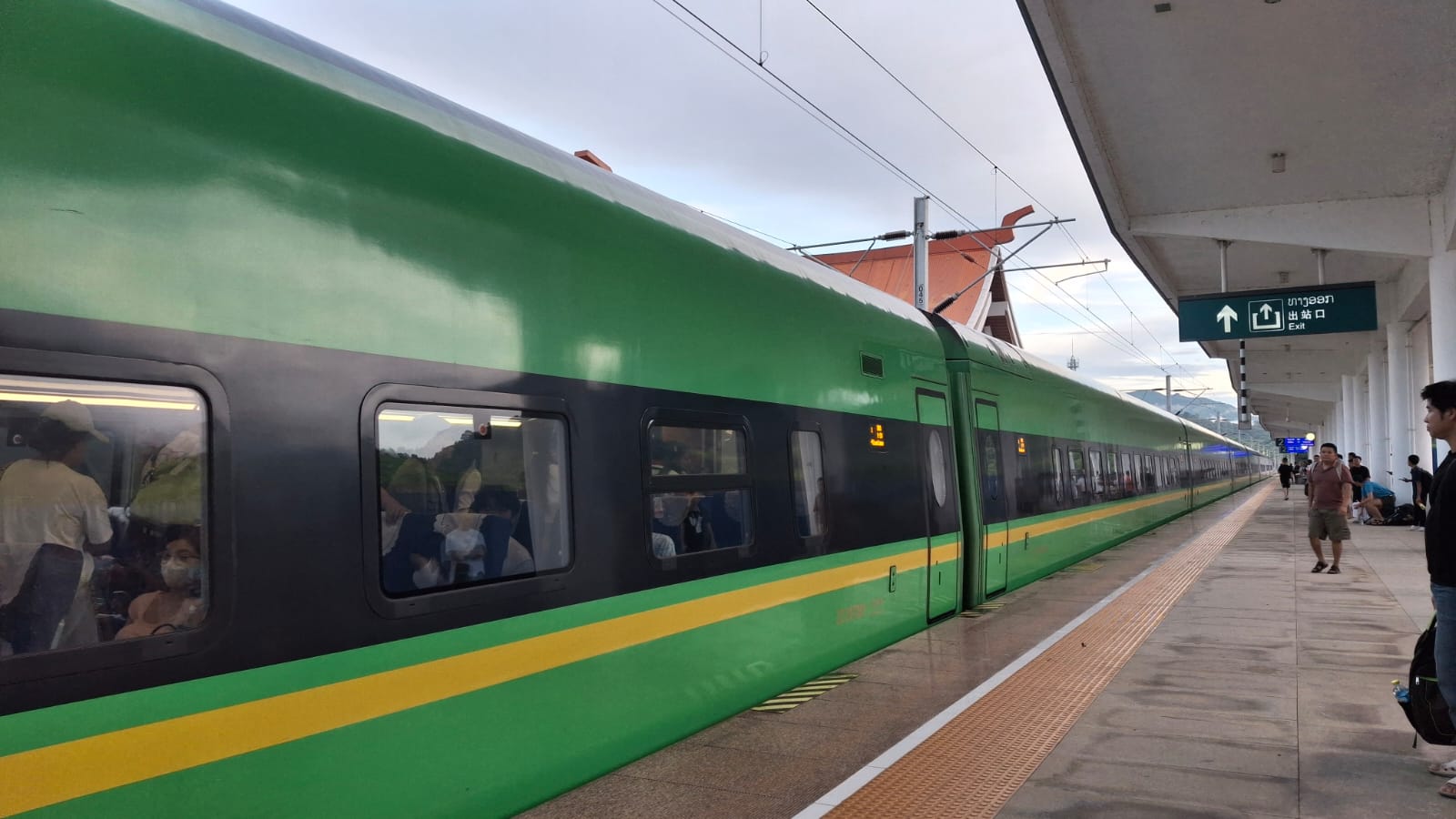 Rame verte et jaune du train Laos-Chine à quai en gare de Luang Prabang, voyageurs à bord et panneau de sortie en lao
